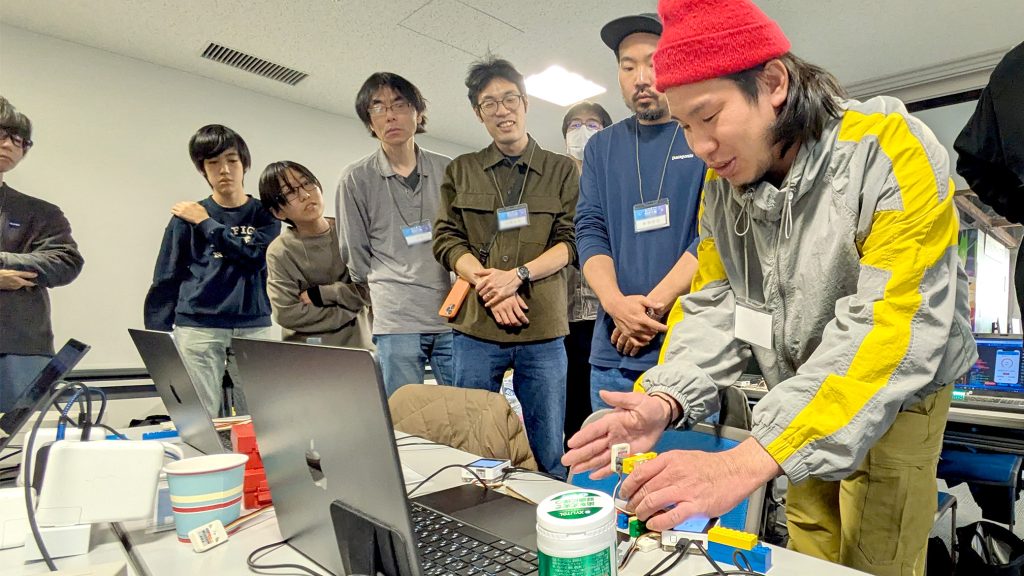 2日目、成果発表。テルミンのように距離で音程を変えるプロトタイプのデモを参加者らが注目している場面。